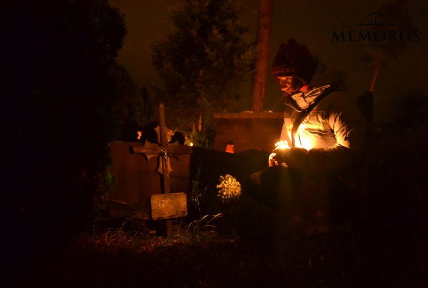 Prie kapavietės tamsoje sėdinti moteris laikanti rankoje žvakę